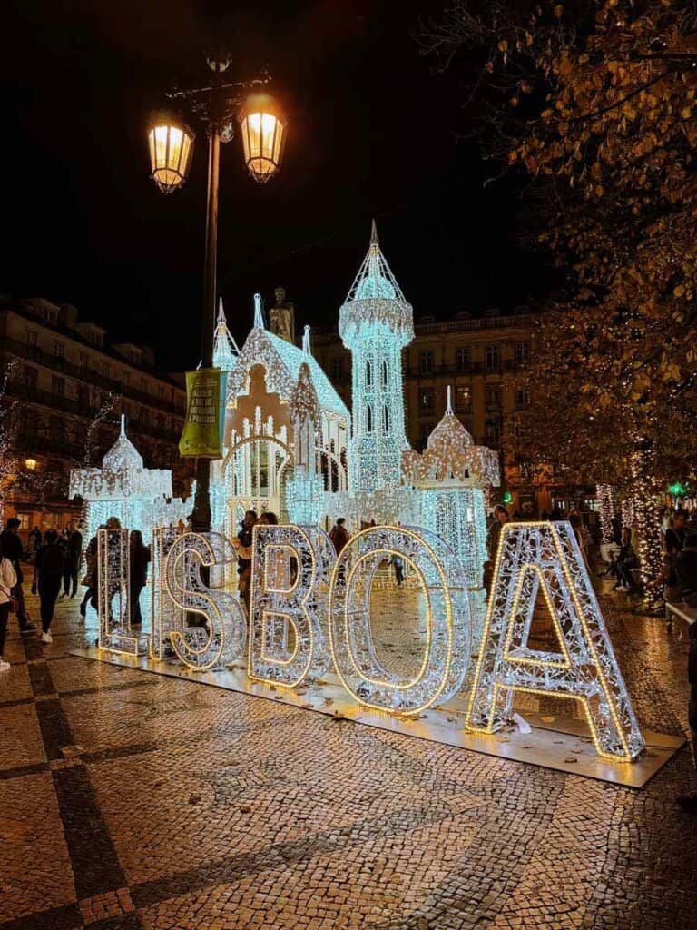 Bola de Natal gigante coberta de milhares de luzes azuis com uma entrada em forma de arco e a palavra "LISBOA" iluminada em primeiro plano. A decoração de Natal em Lisboa destaca-se à noite numa praça com calçada portuguesa enquanto várias pessoas passeiam junto à estrutura luminosa.