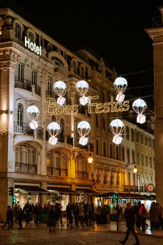 Iluminação de Natal em Lisboa com a mensagem "Boas Festas" em luzes douradas e decorações suspensas em forma de balões de ar quente com presentes. Pessoas passeiam à noite entre os edifícios históricos da Baixa de Lisboa.