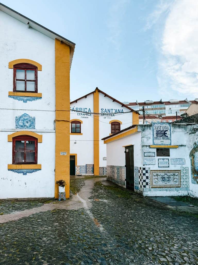The white and mustard yellow exterior of Fábrica Sant'Anna tile factory in Lisbon surrounds a cobbled courtyard with blue and white ceramic tile panels decorating the walls. Large painted lettering reads "FÁBRICA SANT'ANNA CERÂMICA ARTÍSTICA FUNDADA EM 1741".