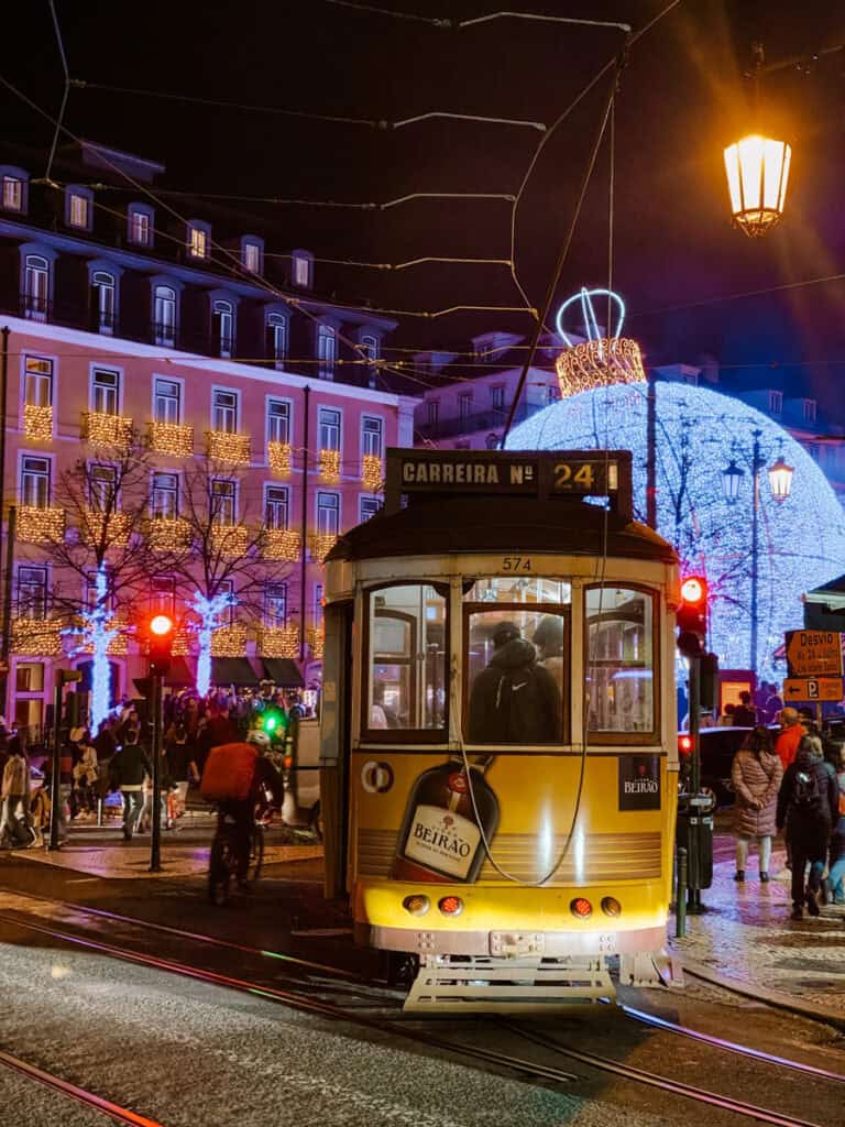 Elétrico amarelo da carreira 24 visto de trás numa rua de Lisboa à noite. Ao fundo destaca-se a iluminação de Natal em Lisboa com uma grande bola azul brilhante e edifícios decorados com luzes douradas.