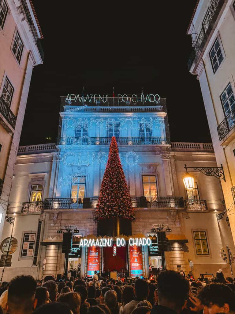 Fachada dos Armazéns do Chiado iluminada com luzes azuis e uma grande árvore de Natal vermelha suspensa sobre a entrada. Uma multidão reúne-se à noite para assistir às celebrações natalícias no centro de Lisboa.