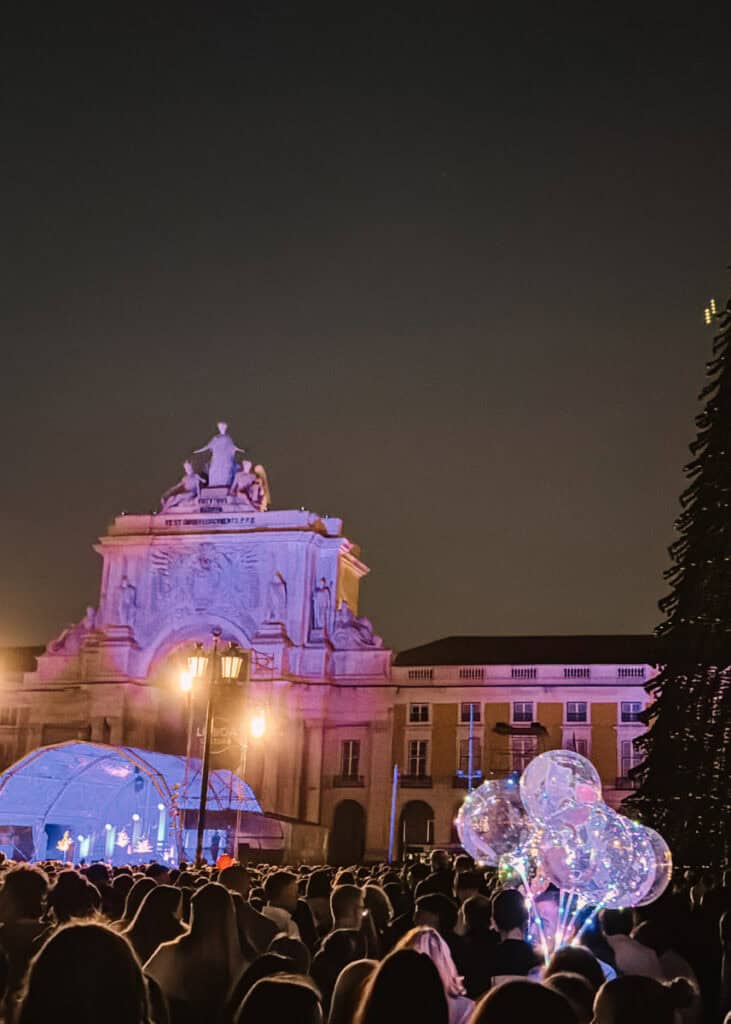 Multidão reunida na Praça do Comércio à noite junto ao Arco da Rua Augusta iluminado em tons de rosa e roxo. Em primeiro plano destacam-se balões transparentes com luzes LED e ao fundo uma estrutura luminosa azul de Natal.