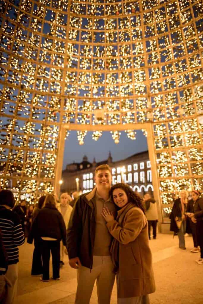 Casal sorridente abraçado durante uma sessão de fotos de Natal em Lisboa, no interior de uma árvore de Natal gigante iluminada com milhares de luzes douradas. Ao fundo veem-se outras pessoas e os edifícios históricos da Praça do Comércio ao anoitecer.