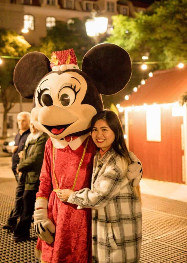 Mulher sorridente com um casaco aos quadrados abraçada a uma personagem vestida de Minnie Mouse com um vestido vermelho e um chapéu natalício. Ao fundo veem-se luzes e uma banca de Natal iluminada numa rua de Lisboa durante a noite.