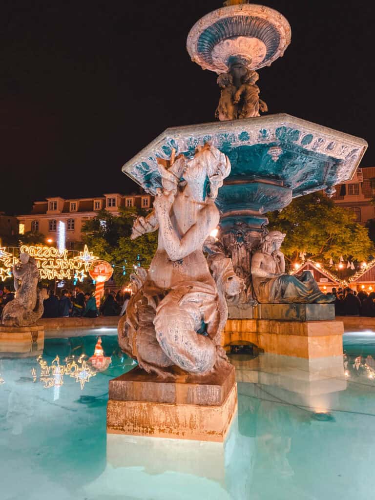 Fonte monumental da Praça do Rossio em Lisboa iluminada à noite com esculturas de figuras mitológicas e água azul turquesa. Ao fundo destacam-se as luzes de Natal em Lisboa com estrelas douradas e bancas do mercado natalício rodeadas de visitantes.