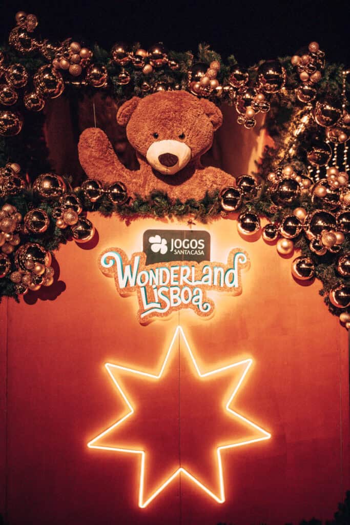 Urso de peluche castanho gigante rodeado por grinaldas verdes e bolas de Natal douradas no Wonderland Lisboa. Por baixo destacam-se uma estrela luminosa e os letreiros "Jogos Santa Casa" e "Wonderland Lisboa" numa decoração festiva durante o Natal em Lisboa.
