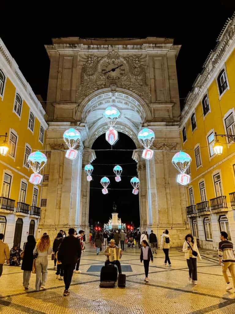 Arco da Rua Augusta iluminado à noite com luzes de Natal em Lisboa em forma de balões coloridos suspensos. Pessoas passeiam pela Rua Augusta enquanto um músico toca acordeão junto ao arco.