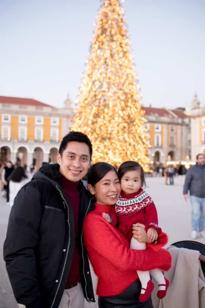Família com uma criança pequena ao colo da mãe a posar sorridente para uma sessão de fotos de Natal na Praça do Comércio em Lisboa. Ao fundo destaca-se uma árvore de Natal gigante iluminada com luzes douradas entre os edifícios históricos da praça.