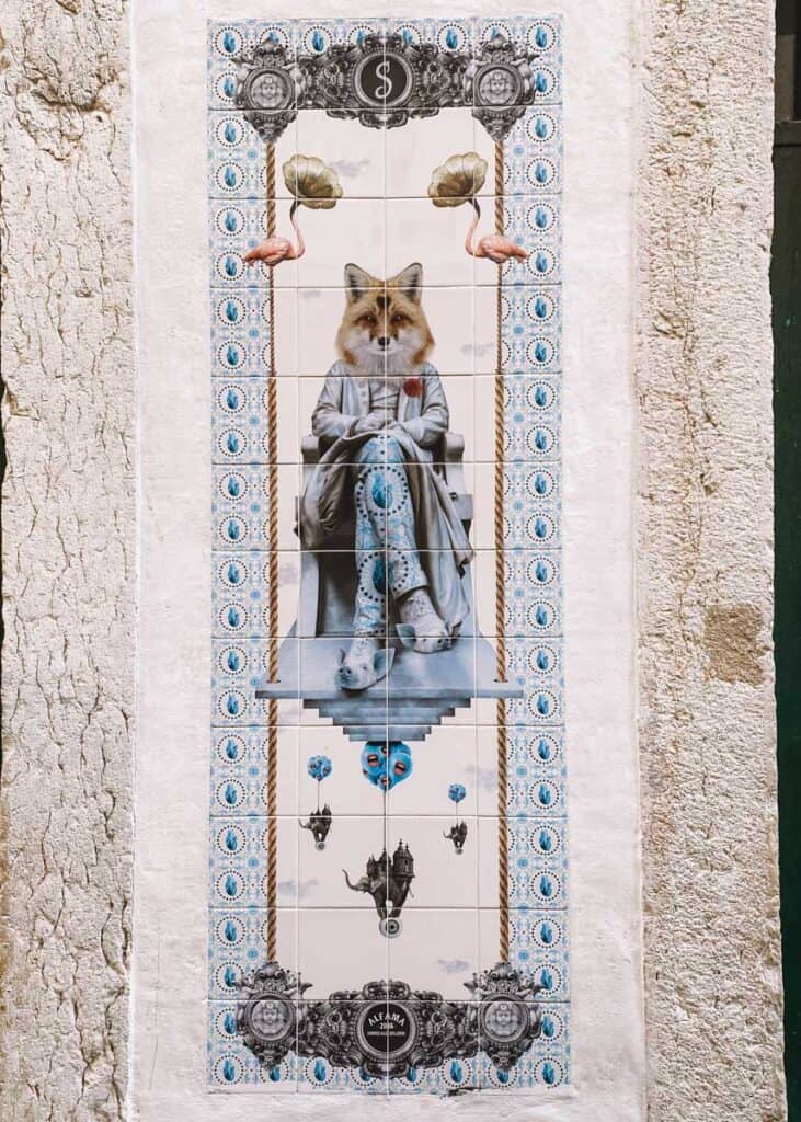 A tall Surrealejos ceramic tile mural in Lisbon shows a fox headed figure dressed in formal clothing seated on a suspended chair. Flamingos blue patterned borders floating masks and other surreal details surround the central figure.