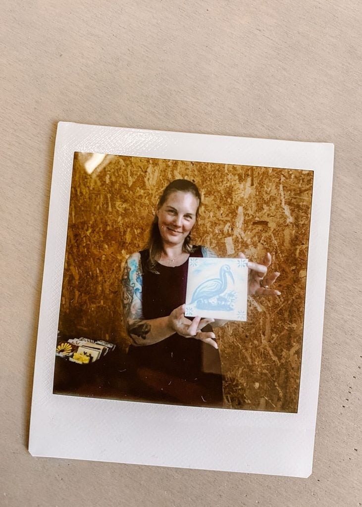 A Polaroid photograph shows a smiling woman holding her finished white ceramic tile painted with a pale blue bird and floral corner details. The finished piece marks the end of a tile painting workshop.