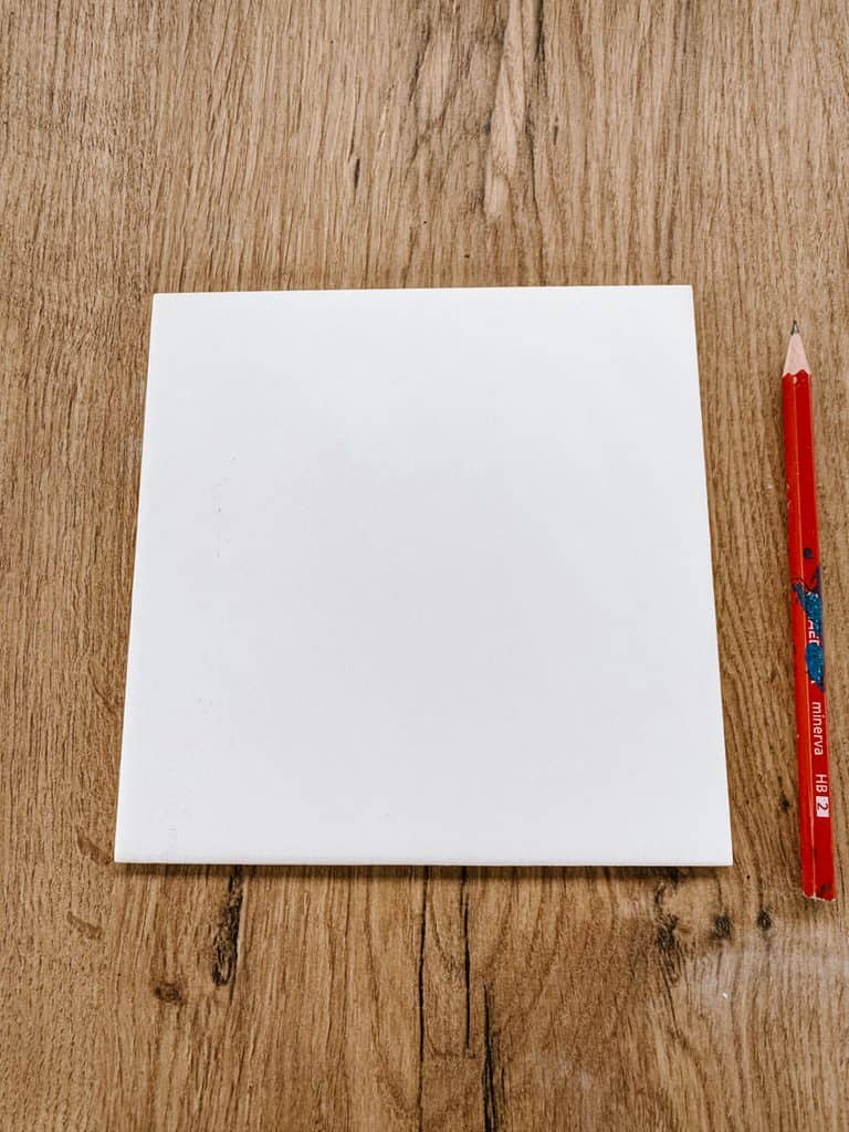 A blank white ceramic tile sits on a wooden table beside a red pencil. The clean tile is ready to be sketched and decorated during a tile painting workshop.