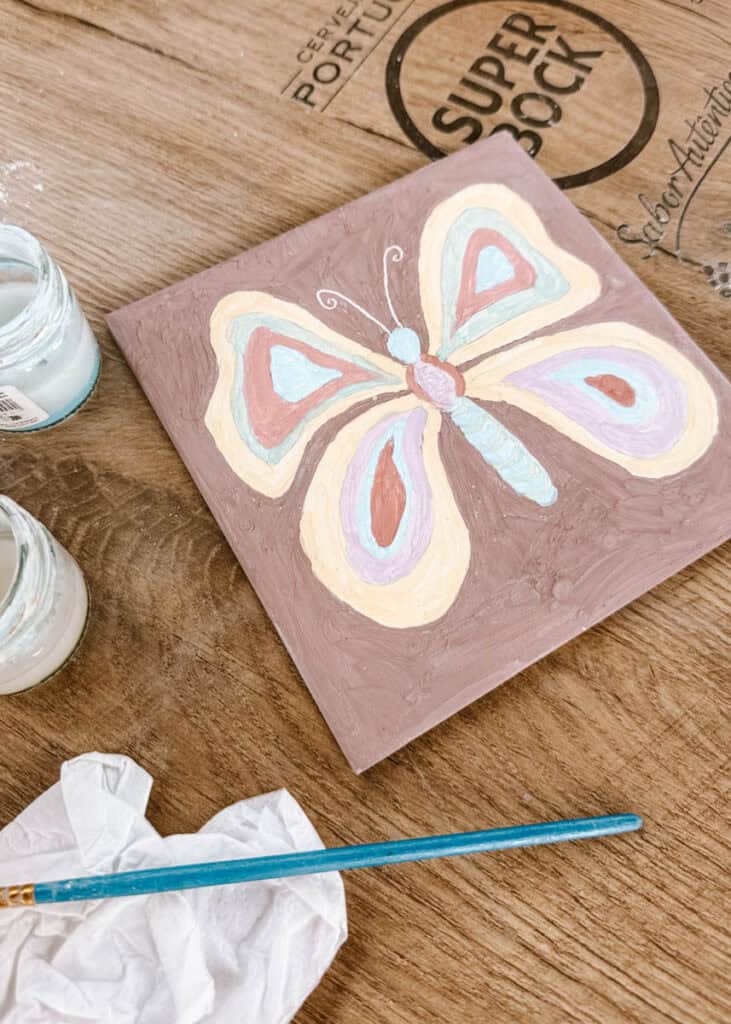 A square ceramic tile painted with a large pastel butterfly in cream blue pink and lilac rests on a wooden table beside open paint jars and a brush. The finished design shows the creative result of tile painting in Lisbon.