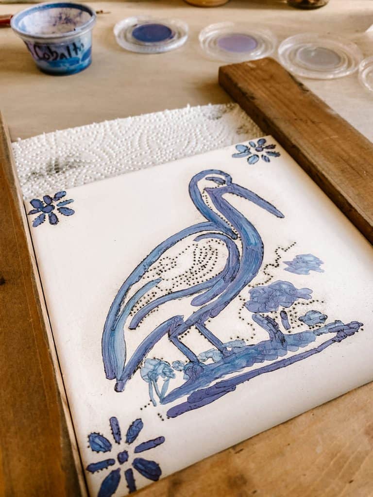 A hand painted ceramic tile shows a large blue and purple bird surrounded by floral details with small blue flowers in each corner. Pots of blue paint sit beside the unfinished design during a tile painting workshop in Lisbon.