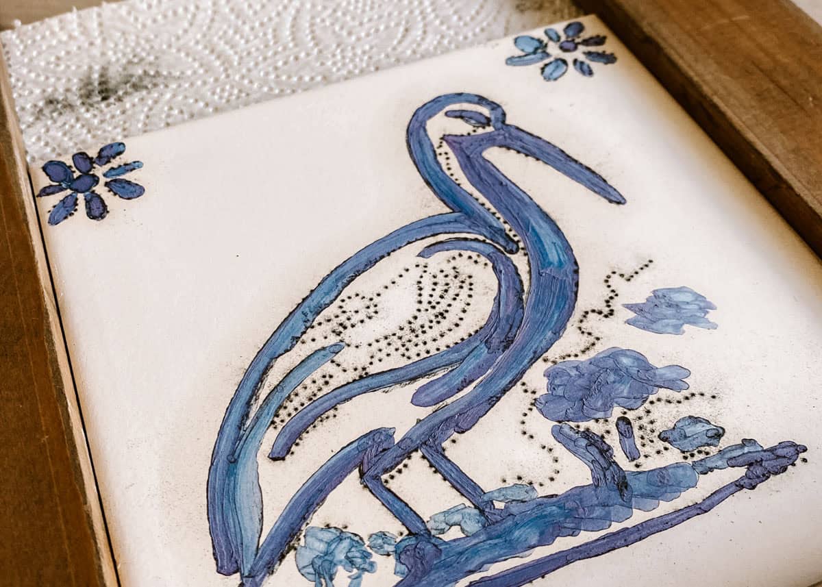 A hand painted ceramic tile decorated with a tall blue bird and small blue floral details. The finished design rests on a wooden work surface during a tile workshop in Lisbon.