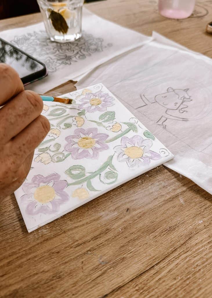 A person paints pale purple flowers with yellow centres and green stems onto a white ceramic tile. Tracing paper with other designs rests beside the tile during a tiles workshop in Lisbon.