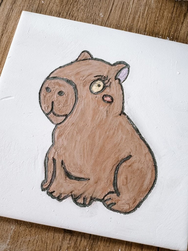 A white ceramic tile painted with a brown capybara sitting upright with a rounded snout and pink cheek. The playful animal design was created during a tiles workshop in Lisbon.