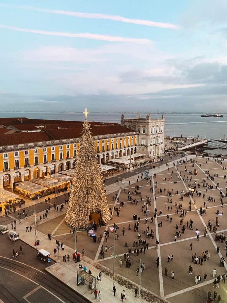 Vista panorâmica da Praça do Comércio em Lisboa ao entardecer com uma árvore de Natal gigante iluminada e várias pessoas a passear. As luzes de Natal em Lisboa decoram também os mercados natalícios instalados junto aos edifícios amarelos da praça com o rio Tejo ao fundo.
