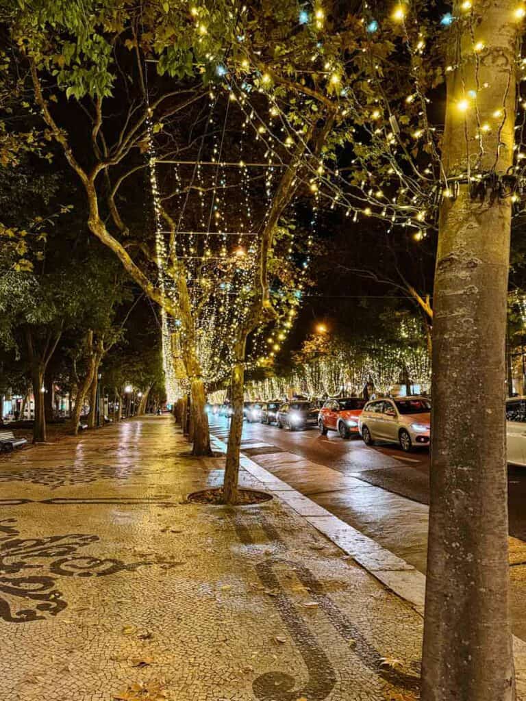 Árvores Na avenida da liberdade em Lisboa decoradas com milhares de luzes de Natal douradas e azuis que formam um túnel luminoso sobre o passeio de calçada portuguesa. Ao lado, vários carros circulam pela estrada enquanto as luzes natalícias iluminam a noite.