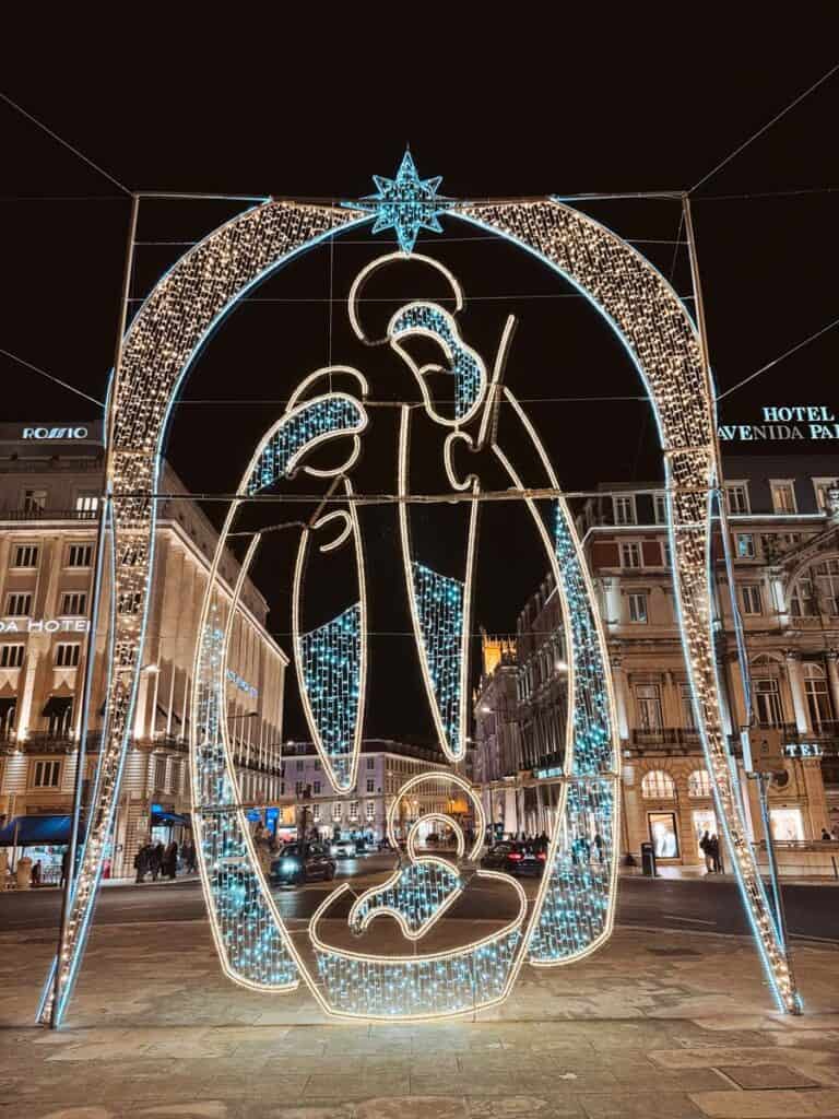 Estrutura luminosa em forma de arco com um presépio estilizado em luzes brancas e azuis no centro de uma praça de Lisboa à noite. Esta decoração de Natal em Lisboa enquadra a rua ao fundo com carros, edifícios históricos e pessoas a circular pela zona do Rossio.
