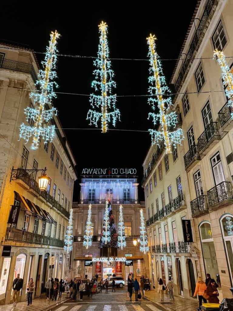 Rua do Carmo em Lisboa iluminada com decorações de Natal suspensas em forma de árvores luminosas azuis e douradas. Ao fundo destacam-se os Armazéns do Chiado decorados com uma grande árvore de Natal enquanto várias pessoas passeiam pela rua comercial.