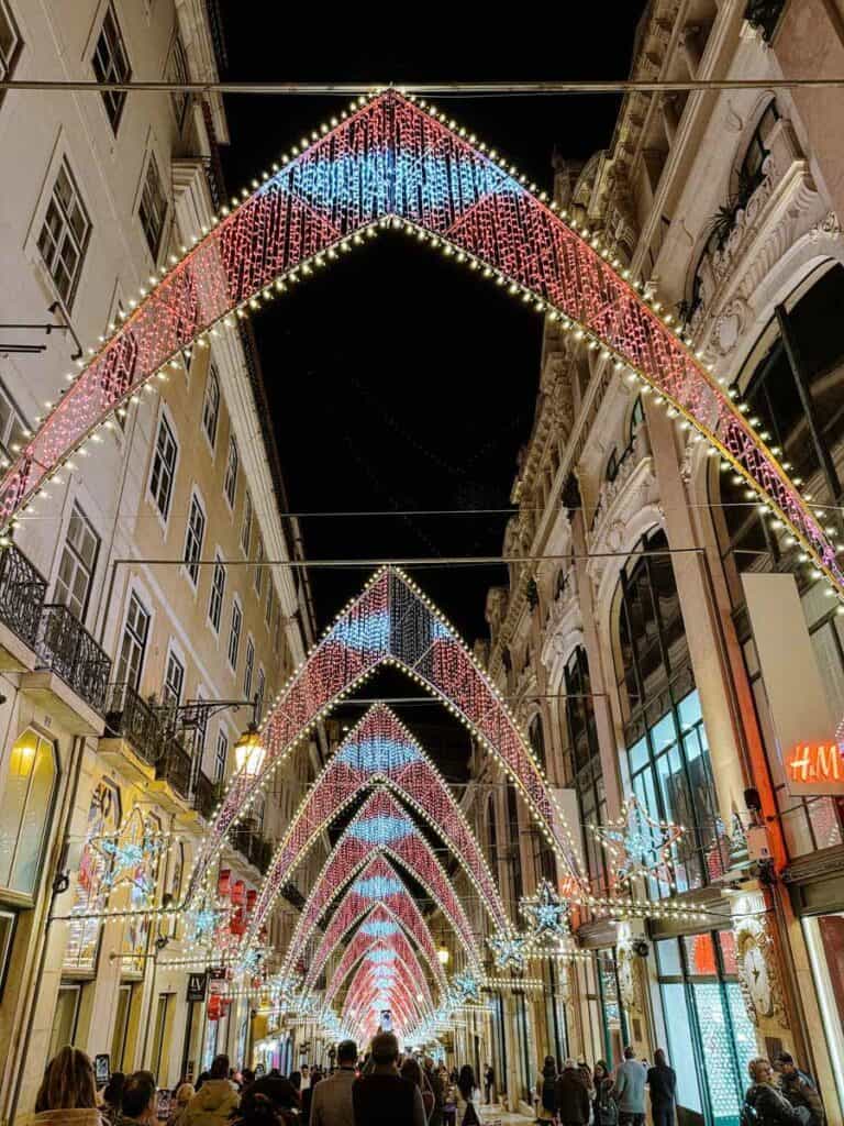 Rua Augusta em Lisboa decorada com arcos de luzes de Natal vermelhas, azuis e douradas que formam um túnel luminoso entre os edifícios históricos. Várias pessoas passeiam à noite sob a iluminação de Natal em Lisboa, acompanhada por estrelas brilhantes suspensas ao longo da rua.