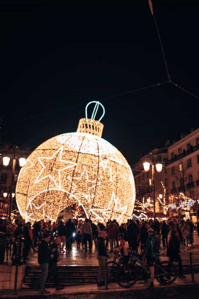 Bola de Natal gigante decorada com luzes douradas e estrelas brancas numa praça de Lisboa à noite. Uma multidão passeia à volta da estrutura luminosa durante as celebrações do Natal em Lisboa.