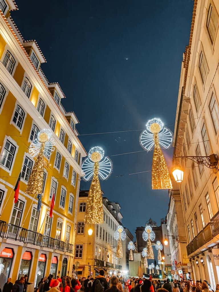 Rua Augusta em Lisboa decorada com anjos de Natal suspensos entre os edifícios históricos, iluminados com luzes brancas e douradas. Uma multidão passeia ao anoitecer sob as luzes de Natal em Lisboa, com o Arco da Rua Augusta visível ao fundo.