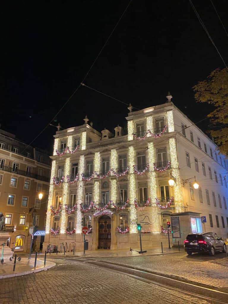 Edifício de esquina em Lisboa decorado com cortinas de luzes douradas e grinaldas vermelhas e brancas ao longo das varandas. As luzes de Natal em Lisboa destacam a fachada histórica durante a noite, junto a uma rua de calçada com carros e candeeiros acesos.
