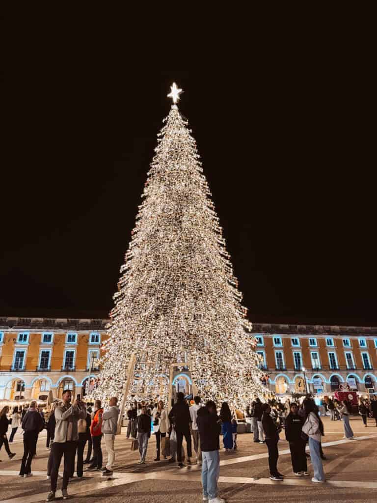 Árvore de Natal gigante iluminada com milhares de luzes douradas e uma estrela brilhante no topo na Praça do Comércio em Lisboa. Várias pessoas passeiam e tiram fotografias junto à árvore que se destaca na decoração de Natal em Lisboa.