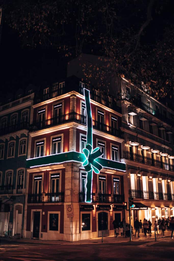 Edifício de esquina em Lisboa decorado com fitas luminosas verdes e brancas e um grande laço de Natal na fachada. A iluminação natalícia destaca-se à noite entre os edifícios históricos enquanto várias pessoas passeiam pela rua.
