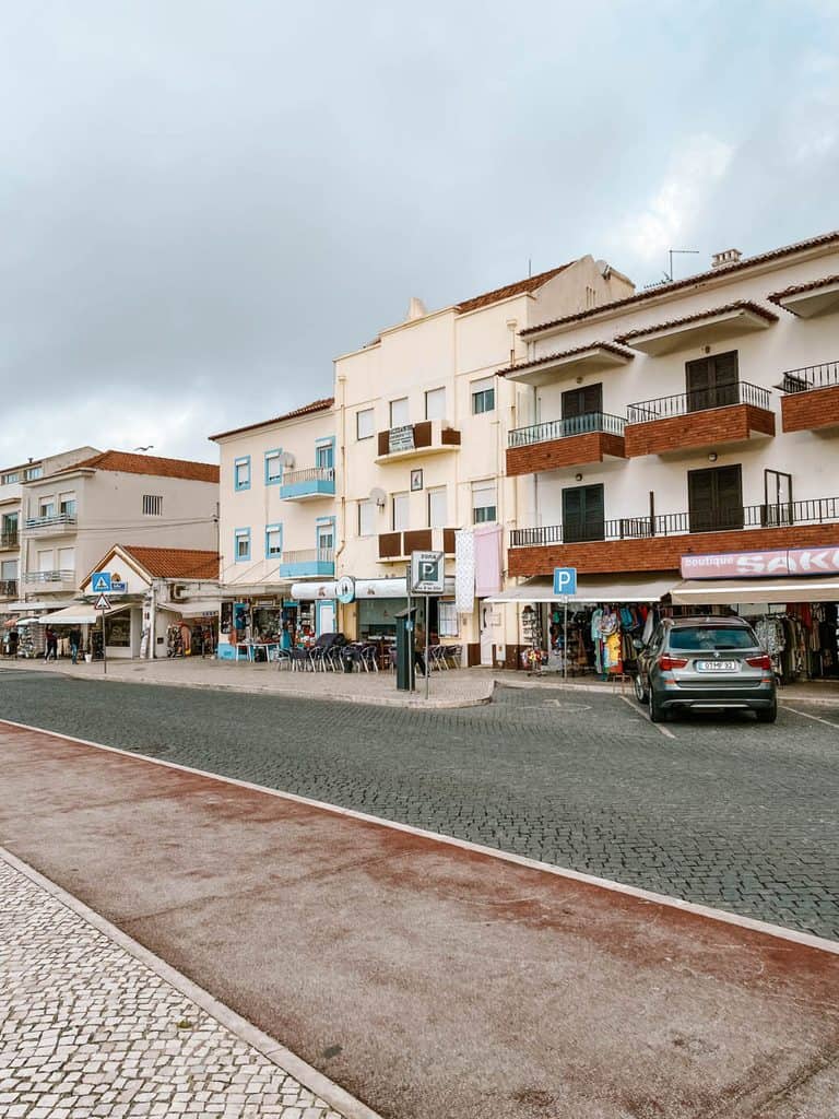 Boulevard in Nazaré met witte en crèmekleurige huizen met balkons en roodbruine dakpannen. Op de begane grond zitten kleine winkeltjes en cafés met terrasjes langs een straat met donkere klinkers.