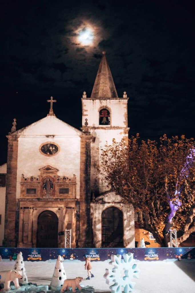 Pista de patinagem da Vila Natal de Óbidos com figuras de ursos polares e flocos de neve iluminados diante de uma igreja histórica. A torre sineira destaca-se sob a lua parcialmente escondida pelas nuvens neste cenário noturno de um mercado de Natal em Portugal.