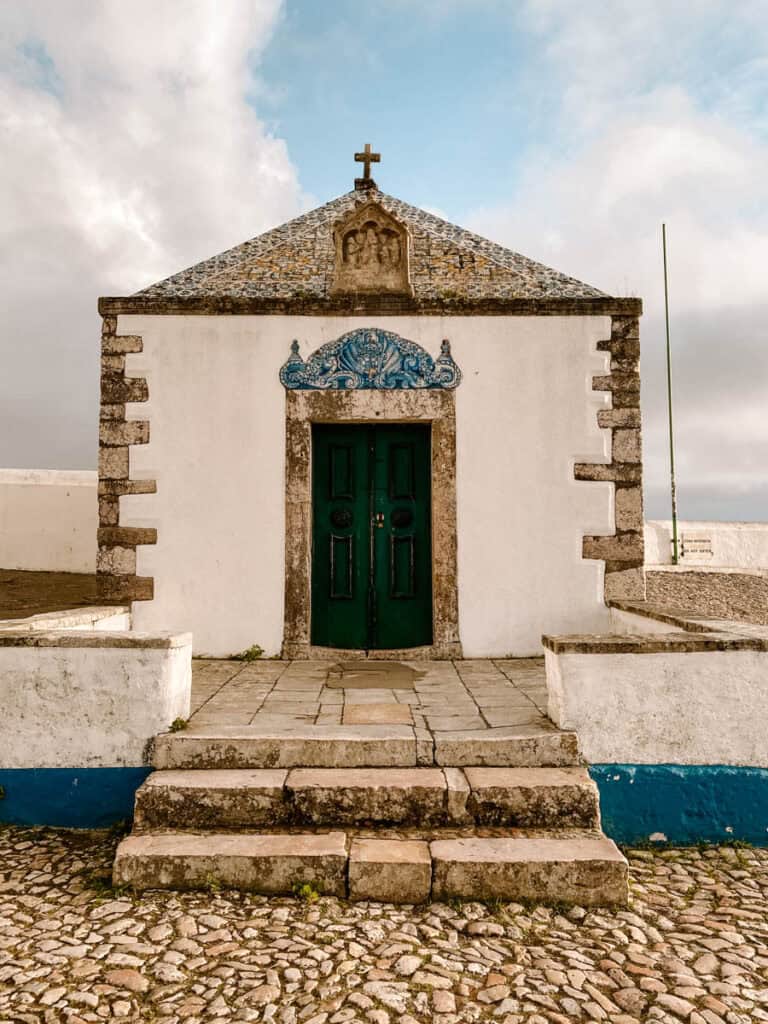 De Ermida da Memória in Nazaré is een kleine witte kapel met een donkergroene deur en blauw versierde tegels boven de ingang. Een stenen trap leidt naar de historische kapel met een kruis op het dak in de wijk Sítio.