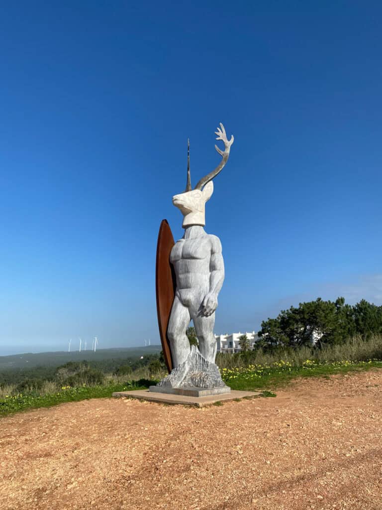 De Escultura Veado Surfista in Nazaré is een opvallend standbeeld van een gespierde mensfiguur met een hertenkop en een groot gewei. Naast het zilvergrijze beeld staat een bruine surfplank en op de achtergrond zie je groene heuvels onder een helderblauwe lucht.