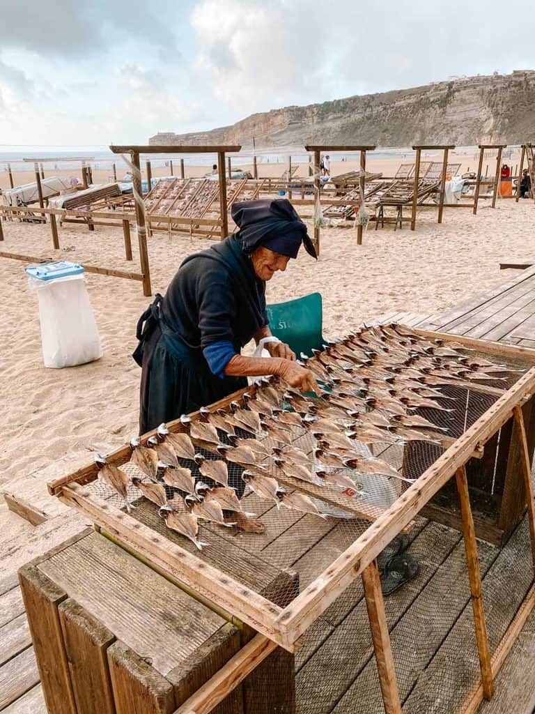 Een oudere vrouw in traditionele zwarte kleding legt vissen te drogen op een houten rek op Praia da Nazaré. Achter haar staan meerdere droogrekken op het zandstrand met de hoge kliffen van Nazaré op de achtergrond.