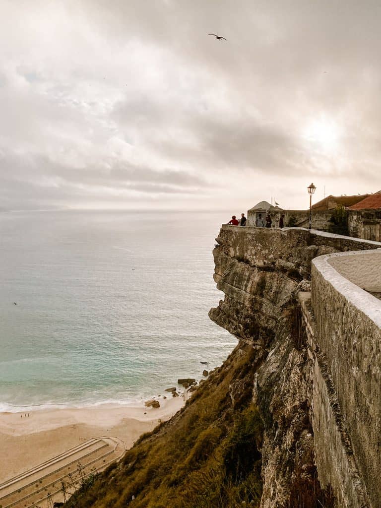 Uitzichtpunt op een hoge rotsklif in Nazaré waar bezoekers uitkijken over de Atlantische Oceaan en het strand beneden. Een stenen muur loopt langs de steile klif onder een bewolkte lucht en laat zien waarom de uitzichtpunten populaire bezienswaardigheden in Nazaré zijn.