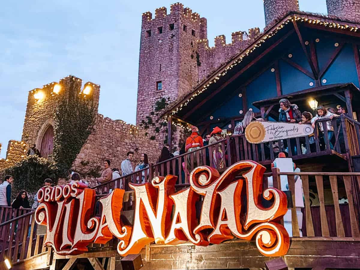 A large illuminated "Óbidos Vila Natal" sign stands in front of the medieval Óbidos Castle in Portugal. Visitors gather on a wooden terrace outside the toy factory during the Óbidos Christmas Village festivities with twinkling lights decorating the castle walls and roofline.