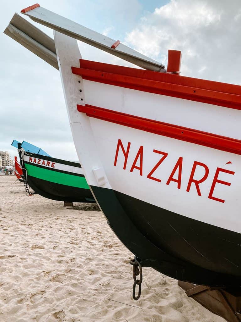 Twee traditionele Portugese vissersboten staan op het zand van Praia da Nazaré. De voorste boot is wit met rode en zwarte details en draagt de tekst "NAZARÉ" in grote rode letters.