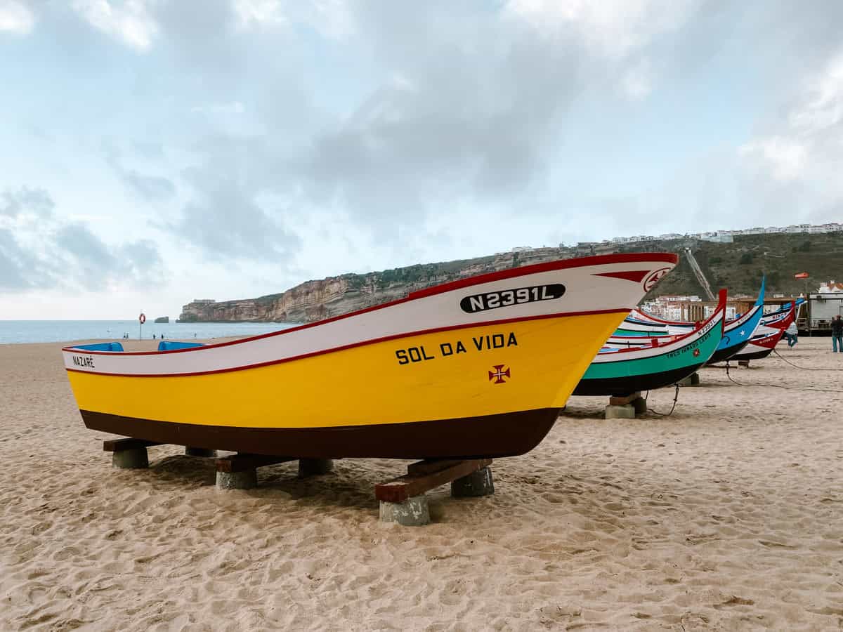 Things to do in Nazaré Portugal include seeing the colourful traditional fishing boats lined up along the sandy beach. A large yellow boat named Sol da Vida sits in the foreground with more brightly painted boats behind it and coastal cliffs in the distance.