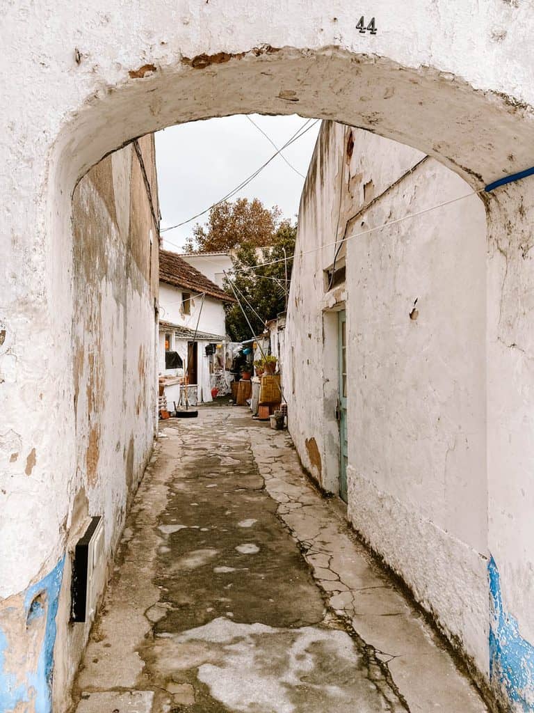 A narrow passageway beneath a weathered whitewashed archway marked with the number 44. Cracked stone paving leads between old Portuguese houses with peeling plaster walls and faded blue paint towards a small courtyard with potted plants.