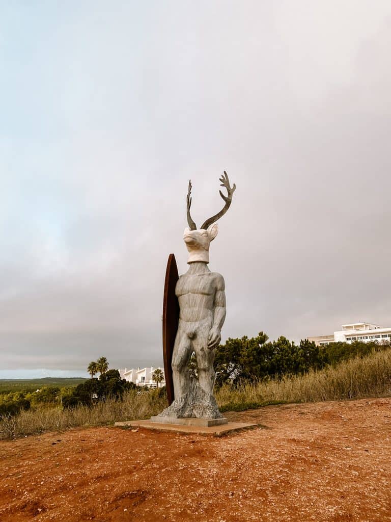 The Escultura Veado Surfista in Nazaré Portugal shows a tall surreal statue with a human body, deer head and antlers, standing beside a surfboard. The sculpture stands on a rocky base in an open grassy area under a cloudy sky with trees and low buildings in the background.