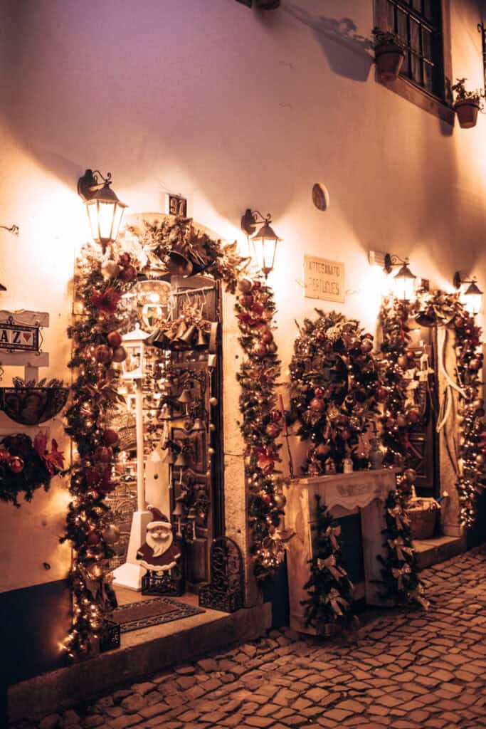Fachada de uma loja de artesanato em Óbidos iluminada à noite, com grinaldas de Natal, bolas vermelhas e douradas, sinos e coroas decorativas à volta da porta e das montras. Um Pai Natal decorativo junto à entrada e os candeeiros de parede criam um ambiente acolhedor durante a época da Vila Natal de Óbidos.