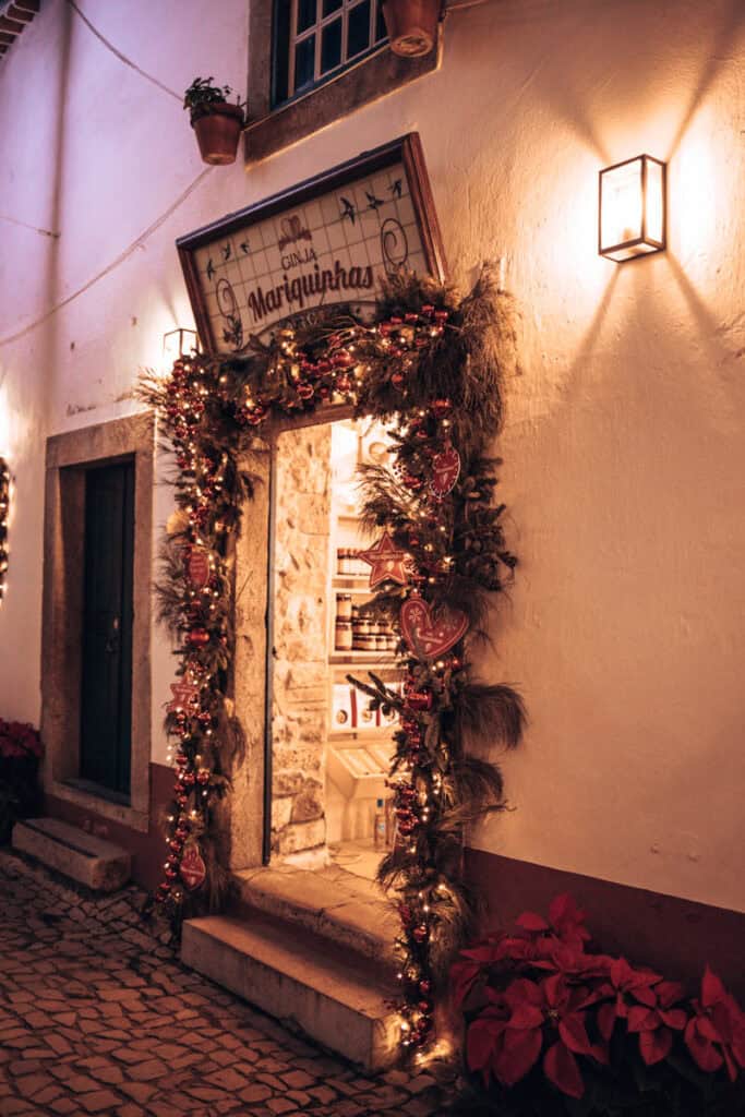 Entrada da loja de ginjinha Mariquinhas em Óbidos, decorada com grinaldas de Natal, luzes douradas, bolas vermelhas e enfeites em forma de coração. A fachada branca e a porta iluminada revelam o interior da loja, criando um ambiente acolhedor durante a época natalícia.