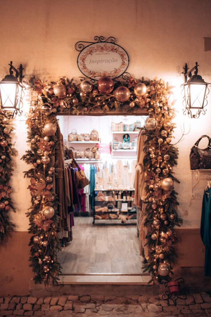 Entrada da loja "Inspiração" em Óbidos decorada com grinaldas iluminadas e bolas de Natal douradas e acobreadas. No interior veem-se roupas e acessórios expostos enquanto os candeeiros de parede e as luzes natalícias criam um ambiente acolhedor.