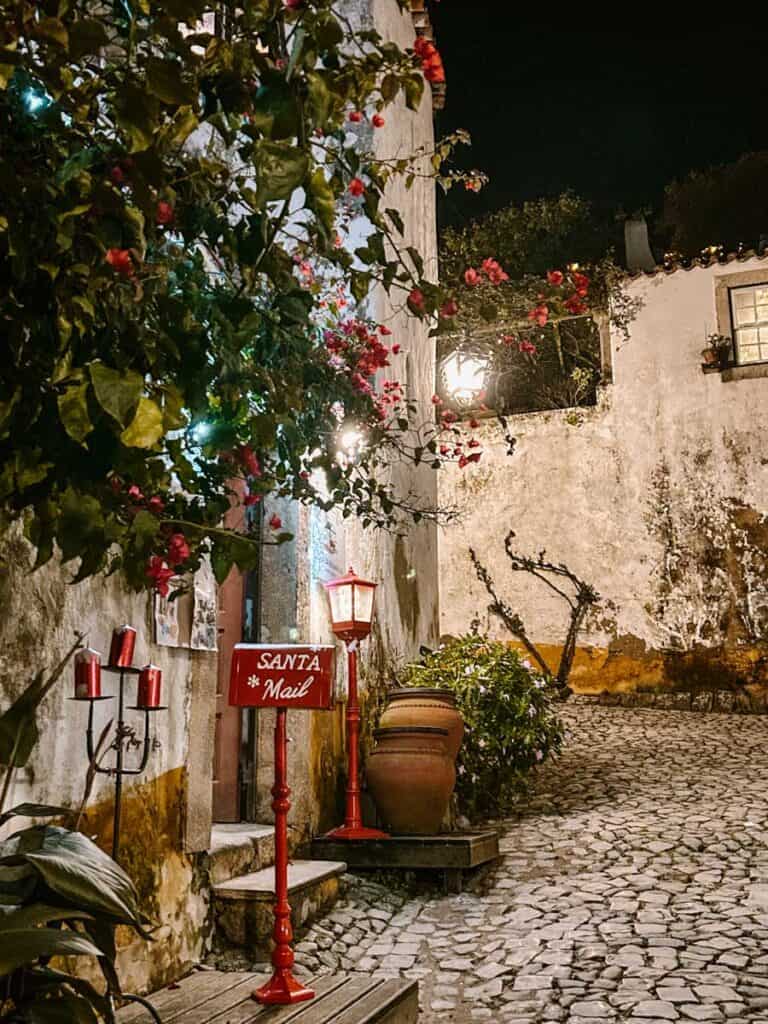 Rua estreita de calçada portuguesa em Óbidos à noite, com casas de paredes brancas, flores vermelhas e iluminação acolhedora. Uma caixa de correio vermelha com a inscrição "SANTA Mail" e um candeeiro decorativo criam um cenário natalício nas ruas da Vila Natal Óbidos 2026