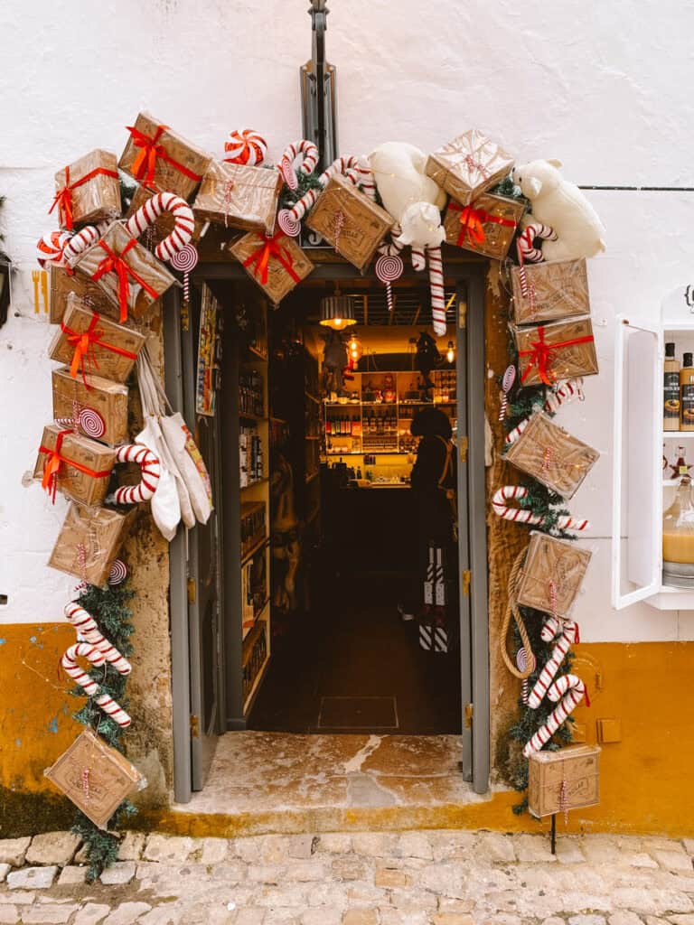 Entrada de uma loja tradicional em Óbidos decorada para o Natal com caixas de presentes, laços vermelhos, bengalas doces, rebuçados às riscas e ursos de peluche brancos. No interior, a iluminação acolhedora revela prateleiras com produtos regionais, criando um ambiente natalício nas ruas da vila.