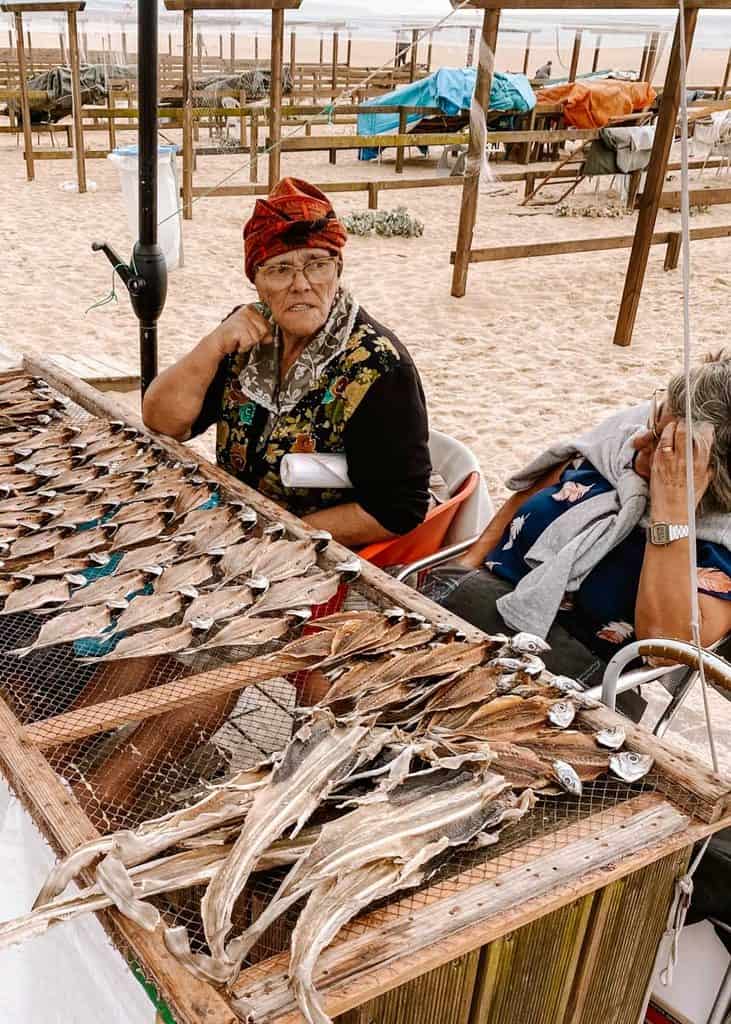 Twee oudere Portugese vrouwen zitten naast een houten droogrek vol opengesneden vissen op het strand van Nazaré. Een van de vrouwen draagt een kleurrijke gebloemde blouse en een rood gestreepte hoofddoek die kenmerkend is voor de traditionele visserscultuur van Nazaré.