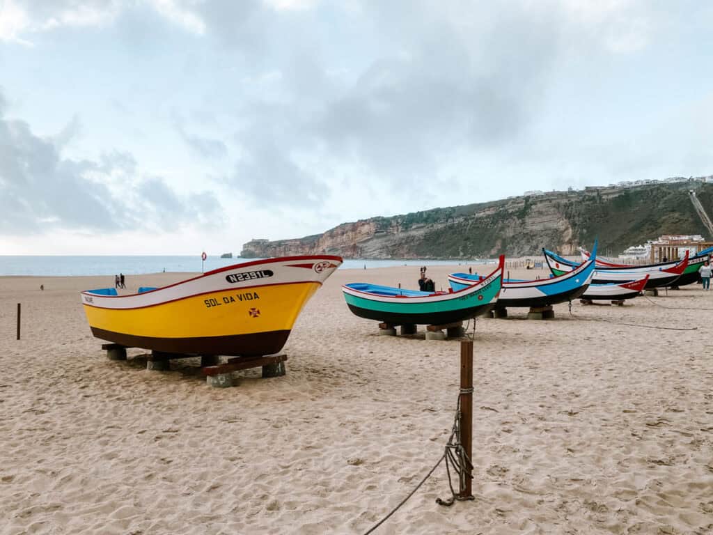 Kleurrijke traditionele vissersboten in geel, groen, blauw en rood staan op het brede zandstrand van Nazaré in Portugal. Op de achtergrond zie je de hoge kliffen langs de kust en de Atlantische Oceaan.