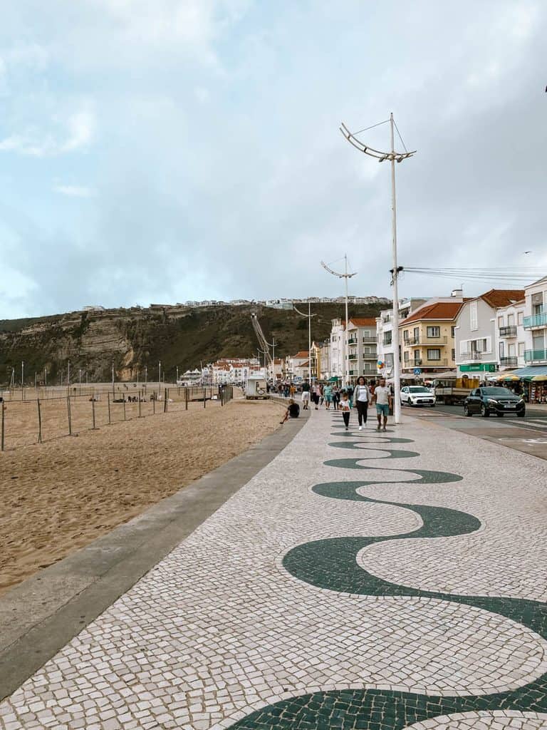 De boulevard langs Praia da Nazaré heeft een mozaïekpatroon van golvende groene lijnen op witte straatstenen. Mensen wandelen langs het brede zandstrand met aan de ene kant kleurrijke huizen en aan de andere kant de hoge kliffen van Nazaré.