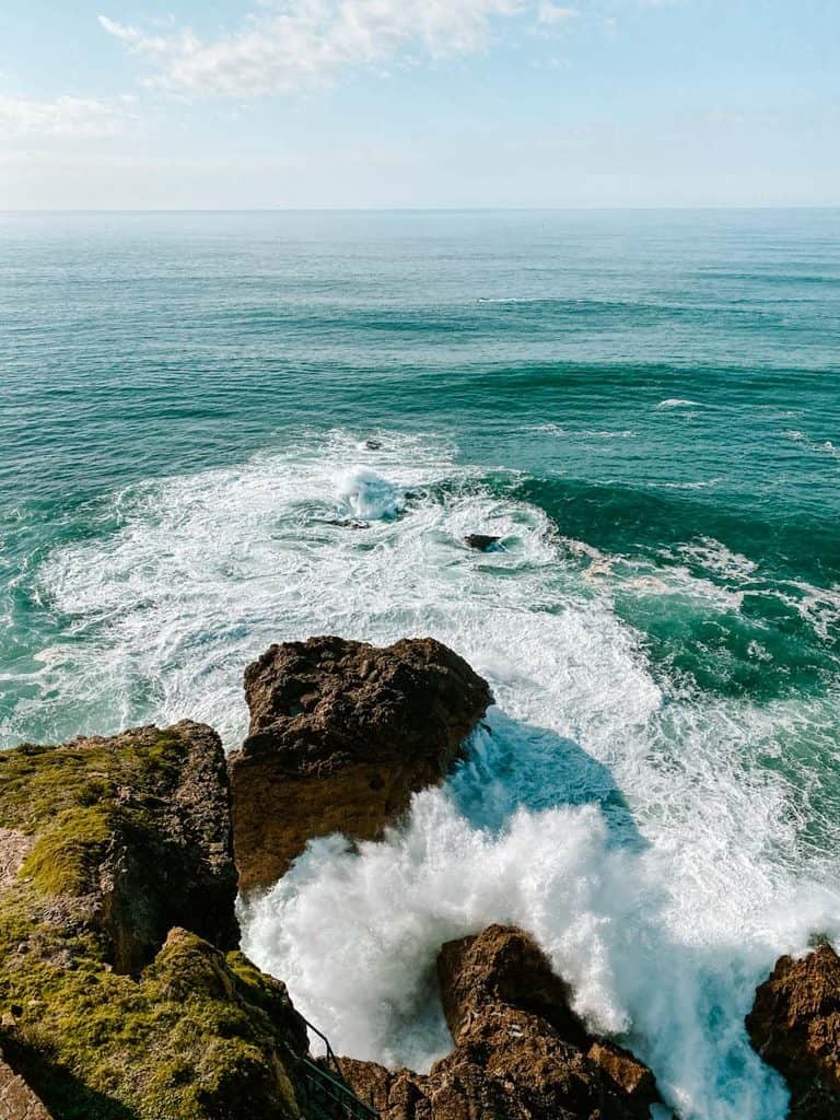 Krachtige golven van de Atlantische Oceaan slaan tegen de donkere rotsen aan de kust van Nazaré. Het helder turquoise zeewater verandert in grote hoeveelheden wit schuim terwijl de golven tussen de met groen mos bedekte rotsen breken.